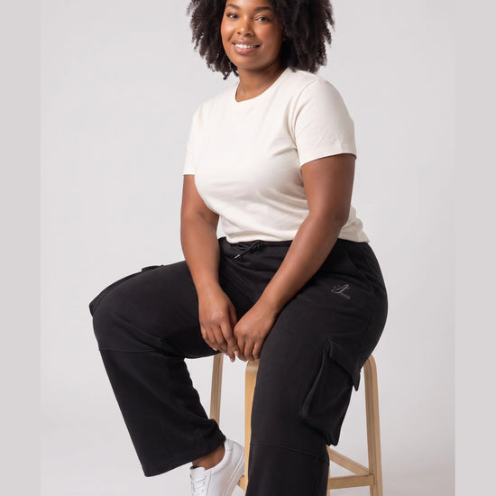Woman seated wearing black Legacy heavyweight organic cotton cargo pants with thigh flap pockets.