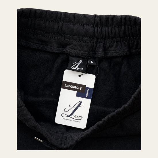 Close-up of black sweatpants with an elastic waistband and a visible Legacy tag, shown against a light beige background.