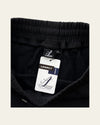 Close-up of black sweatpants with an elastic waistband and a visible Legacy tag, shown against a light beige background.