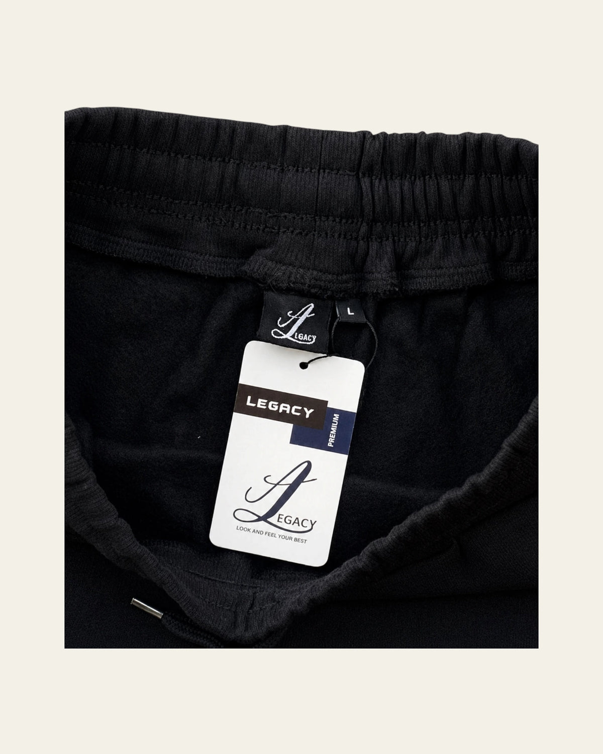 Close-up of black sweatpants with an elastic waistband and a visible Legacy tag, shown against a light beige background.
