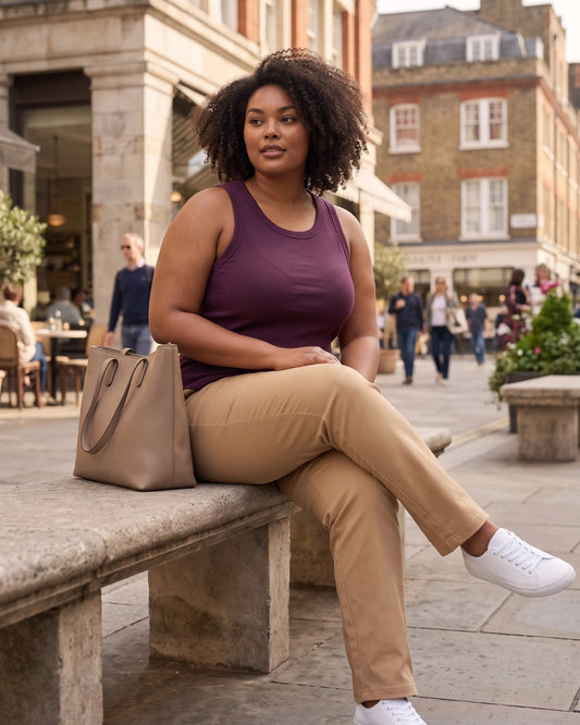 A woman sits on a stone bench in a city plaza wearing a sleeveless plum-purple tank top, beige trousers, and white sneakers, with a large taupe tote bag beside her.